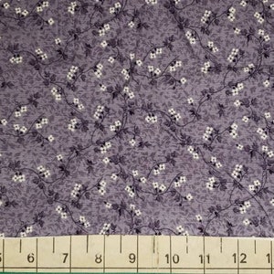 May include: A close-up of a fabric with a floral pattern. The fabric is a muted purple color, with small white flowers and dark gray leaves and stems. A ruler is at the bottom of the image.