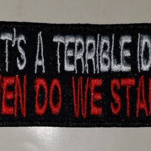 May include: Black rectangular patch with white embroidered text that reads "THAT'S A TERRIBLE IDEA" above red embroidered text that reads "WHEN DO WE START". The patch has a black border.