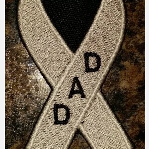 May include: Embroidered ribbon patch in beige with black accents. The patch features the text "RIP" at the top and "DAD" vertically down the center. The background is a mottled brown.