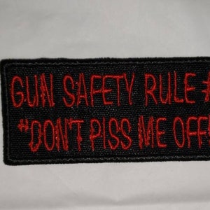 May include: Black rectangular patch with red embroidered text. The text reads "GUN SAFETY RULE #1" and "DON'T PISS ME OFF!". The patch is likely designed to be sewn or attached to clothing or gear.
