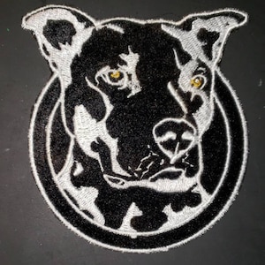 May include: Black and white embroidered patch of a dog's face with a white outline. The dog has a black nose and yellow eyes.