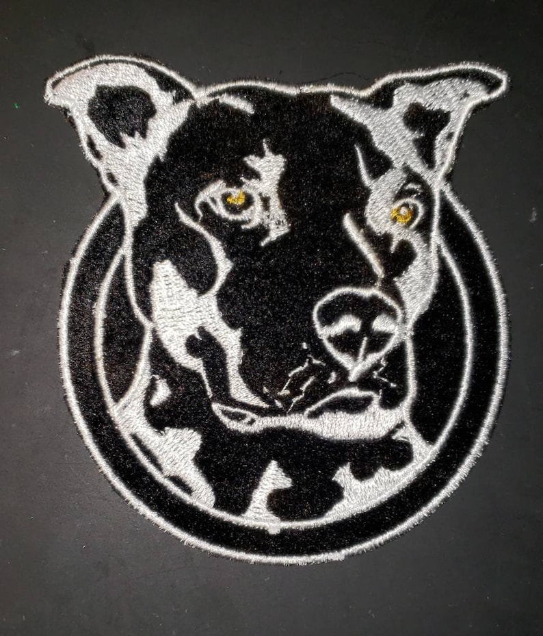 K&ouml;nnte beinhalten: Schwarz-wei&szlig; gestickter Patch mit dem Gesicht eines Hundes mit wei&szlig;em Umriss. Der Hund hat eine schwarze Nase und gelbe Augen.