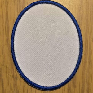 May include: Oval-shaped white patch with a blue stitched border. The patch has a textured surface and is set against a wooden background. This blank patch is suitable for customization.