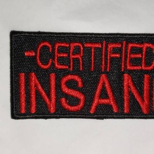 May include: Black rectangular patch with red embroidered text that reads "-CERTIFIED- INSANE-". The patch has a textured appearance and is likely made of fabric. The text is centered and bold.