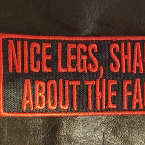 May include: A rectangular patch with the text "NICE LEGS, SHAME ABOUT THE FACE" embroidered in red on a black background. The patch has a red border and is set against a dark, textured surface.