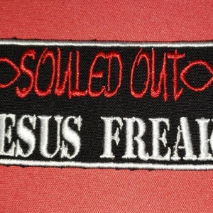 May include: Black and white embroidered patch with red text that reads "SOILED OUT" with a red fish symbol on either side. The bottom of the patch reads "JESUS FREAK" in white.