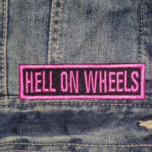 May include: A pink and black embroidered patch with the text "HELL ON WHEELS" on a denim background.