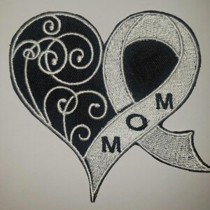 May include: Black and white embroidered patch in the shape of a heart with a silver ribbon. The ribbon is wrapped around the heart and has the word "MOM" embroidered on it.