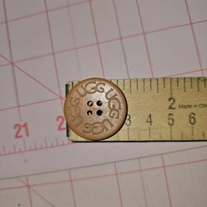 May include: A light brown, round button with the word "UGG" engraved around the edge. The button has four holes in the center. A wooden ruler is next to the button, showing a measurement of approximately 1 inch.