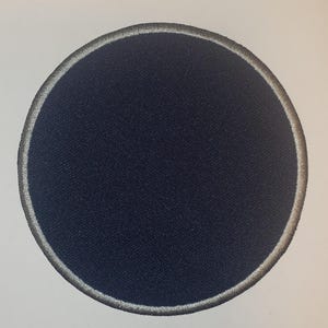 May include: A navy blue, circular patch with a silver embroidered border. The patch has a textured surface and is set against a white background. The design is simple and clean, suitable for various applications.