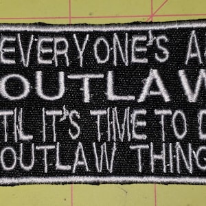 May include: Black embroidered patch with white text that reads "EVERYONE'S AN OUTLAW TIL IT'S TIME TO DO OUTLAW THINGS." The patch has a white border and is rectangular in shape.