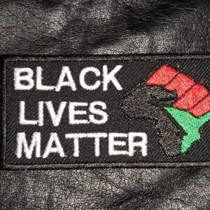 May include: A rectangular black patch with the words "BLACK LIVES MATTER" in white. The patch features a black fist, red knuckles, and a green outline of Africa. The patch is on a black leather surface.