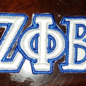 May include: White embroidered letters "Z". "Phi" and "B" with a blue outline. The letters are stitched on a white fabric.
