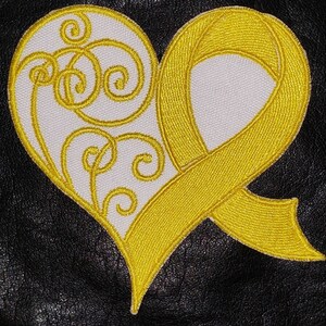 May include: Embroidered patch featuring a heart shape with a white background and gold swirls on the left side. A yellow ribbon is on the right side. The patch is on a black background.