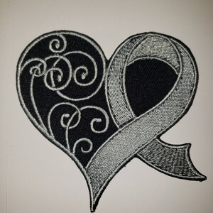 May include: A black and silver embroidered patch in the shape of a heart with a silver ribbon. The heart has a decorative swirl design.
