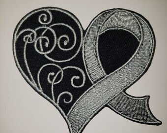 May include: A black and silver embroidered patch in the shape of a heart with a silver ribbon. The heart has a decorative swirl design.
