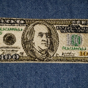 May include: Embroidered patch of a $100 bill featuring a portrait of Benjamin Franklin. The patch is beige with green and gold accents. The text "United States of America" and "100" are visible.