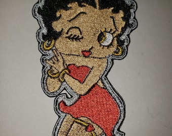 May include: Embroidered patch featuring a cartoon woman winking, wearing a red dress and gold jewelry. The patch has a black outline and a silver border. The woman has black hair and a gold skin tone.