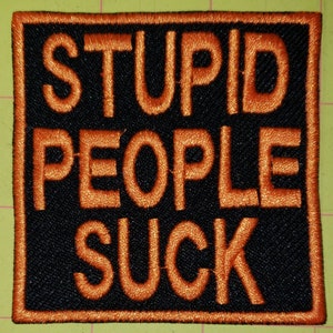 May include: Embroidered patch with the words "STUPID PEOPLE SUCK" in orange thread on a black background. The patch has an orange border and is square-shaped.
