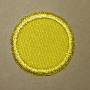 May include: A round, yellow embroidered patch with a textured center and a gold-colored stitched border. The patch is circular in shape and appears to be made of fabric. The background is a neutral tan color.