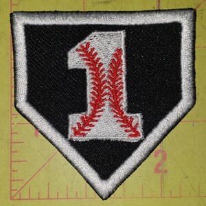 May include: A baseball-themed patch in a home plate shape. The patch features a black background with a white border. A white number "1" is in the center, with red baseball stitching.