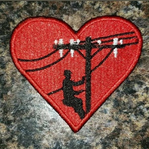 May include: Embroidered red heart patch featuring a black silhouette of a lineman working on a power pole. The patch has a detailed design with black power lines and white insulators, perfect for jackets, bags, or hats.