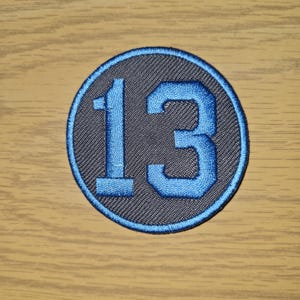 May include: A circular patch with the number 13 embroidered in light blue on a dark gray background. The number is outlined in light blue, and the patch has a light blue border. The patch is on a light brown wooden surface.