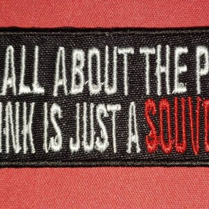 May include: Black embroidered patch with white text that reads "It's all about the pain the ink is just a souvenir" with the word "souvenir" in red.