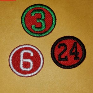 May include: Three circular patches with embroidered numbers. The top patch has a green number 3 on a red background with a green border. The bottom left patch has a white number 6 on a red background with a white border. The bottom right patch has a black number 24 on a red background with a black border.