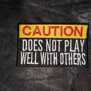 May include: Embroidered patch with the text "CAUTION DOES NOT PLAY WELL WITH OTHERS". The patch has a yellow top border with red text, and white text on a black background. The patch is rectangular and sewn onto a black leather-like surface.