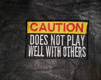 May include: Embroidered patch with the text "CAUTION DOES NOT PLAY WELL WITH OTHERS". The patch has a yellow top border with red text, and white text on a black background. The patch is rectangular and sewn onto a black leather-like surface.