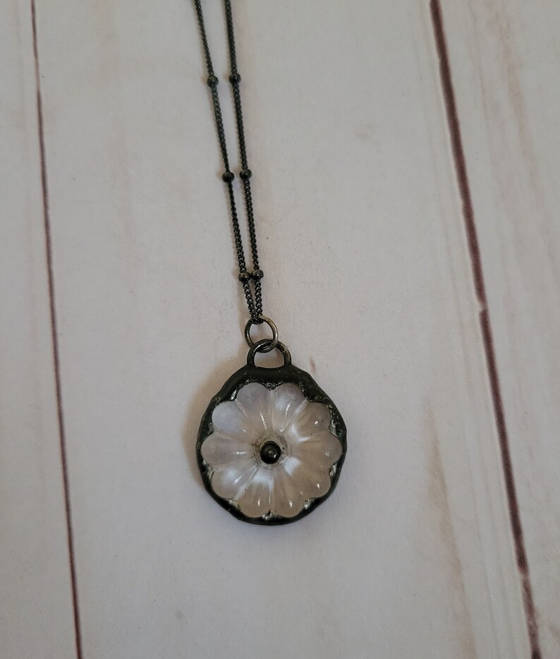 May include: A delicate white flower pendant necklace with a black chain. The flower is made of glass and has a raised design. The chain is made of metal and has a ball chain style.