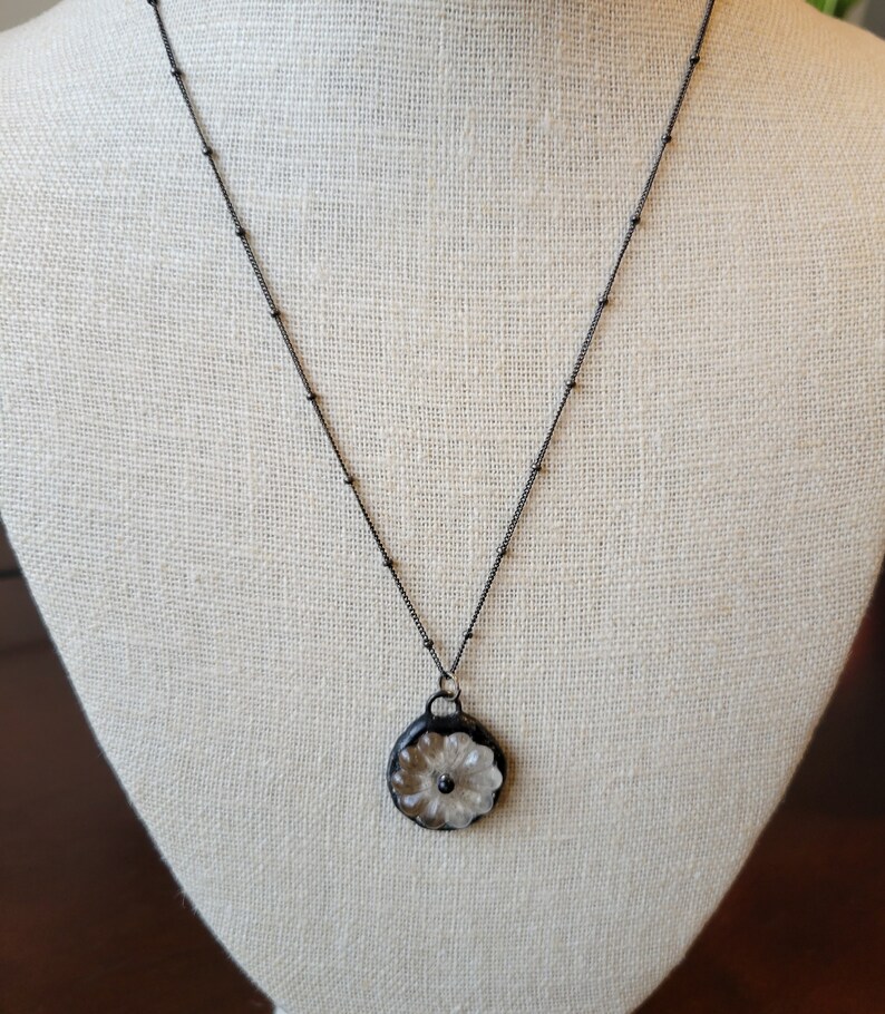 May include: A black chain necklace with a white flower pendant. The flower is carved into a round, clear glass piece.