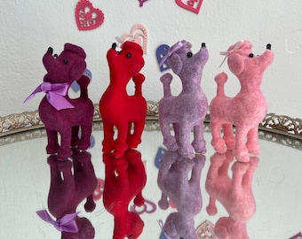 May include: Four plush poodle figurines in shades of purple, red, and pink, each with a bow. They stand on a mirrored surface, reflecting their forms. Decorative paper hearts in various colors are in the background.