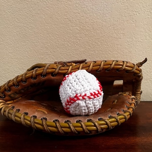 May include: A handmade crochet baseball with red stitching rests inside a vintage leather baseball glove. The glove is brown with tan stitching and a worn appearance, suggesting use. The baseball is white.