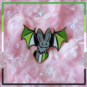 Puede incluir: Un pin de esmalte de murciélago verde, blanco y negro con detalles de purpurina. El murciélago sostiene un pin en forma de corazón con los colores verde, blanco y negro de la bandera del orgullo asexual.