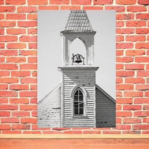 May include: Black and white photograph of a weathered, white church steeple with a bell in the belfry. The steeple has a pointed roof and a single window.