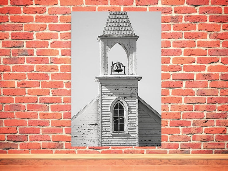 May include: Black and white photograph of a weathered, white church steeple with a bell in the belfry. The steeple has a pointed roof and a single window.