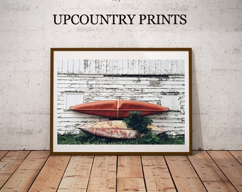 May include: Two canoes, one red and one faded, are hanging on a white weathered wooden wall. The canoes are tied together with rope.