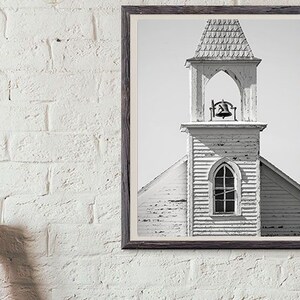 May include: Black and white photograph of a weathered church steeple with a bell in the belfry. The church is made of wood and has a simple design.