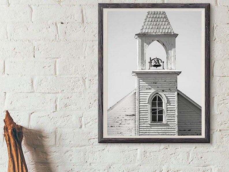 May include: Black and white photograph of a weathered church steeple with a bell in the belfry. The church is made of wood and has a simple design.