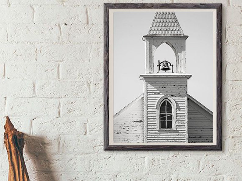 May include: Black and white photograph of a weathered church steeple with a bell in the belfry. The church is made of wood and has a simple design.