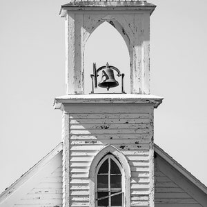 May include: A weathered white church steeple with a bell in the belfry and a window with a pointed arch. The steeple is made of wood and has a simple design.