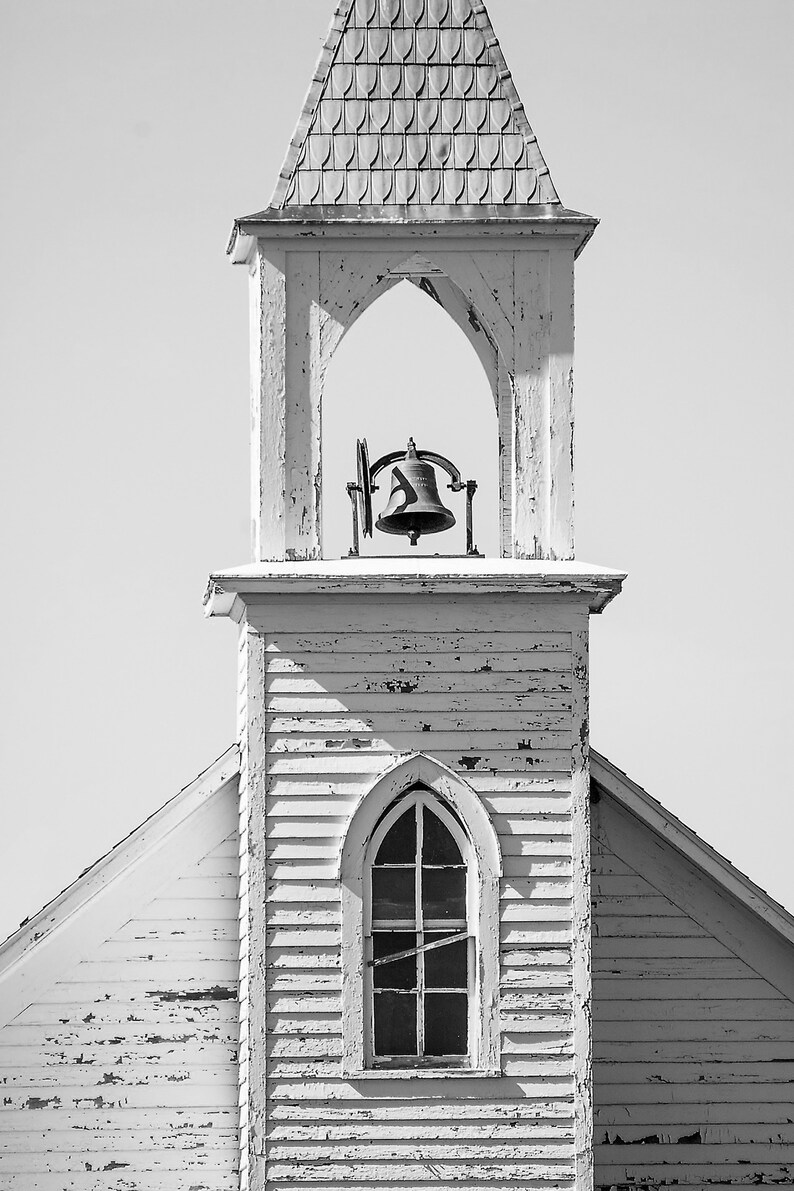 May include: A weathered white church steeple with a bell in the belfry and a window with a pointed arch. The steeple is made of wood and has a simple design.