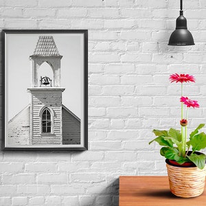 May include: A black and white photograph of a church steeple with a bell in a black frame hanging on a white brick wall. A potted pink gerbera daisy plant sits on a wooden table in front of the picture.