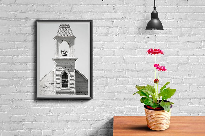 May include: A black and white photograph of a church steeple with a bell in a black frame hanging on a white brick wall. A potted pink gerbera daisy plant sits on a wooden table in front of the picture.
