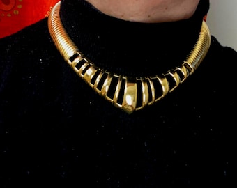 May include: A gold-toned choker necklace with a ribbed design and a V-shaped front. The necklace is worn against a black turtleneck, creating a contrast that highlights the jewelry's metallic sheen. The necklace has a bold, geometric design.