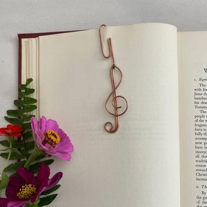 May include: A copper wire bookmark shaped like a treble clef. The bookmark is resting on an open book with a pink flower and green leaves on the left side of the page. The book has text about the significance of Jesus Christ.