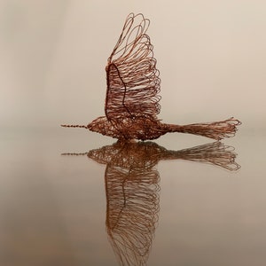 以下が含まれることがあります： 広げた翼で飛んでいる鳥のワイヤースカルプチャー。彫刻は銅線で作られており、鏡のような表面に映っています。