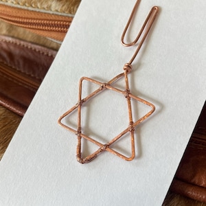 May include: A copper wire star of David pendant hanging from a copper paperclip. The pendant is on a white card.